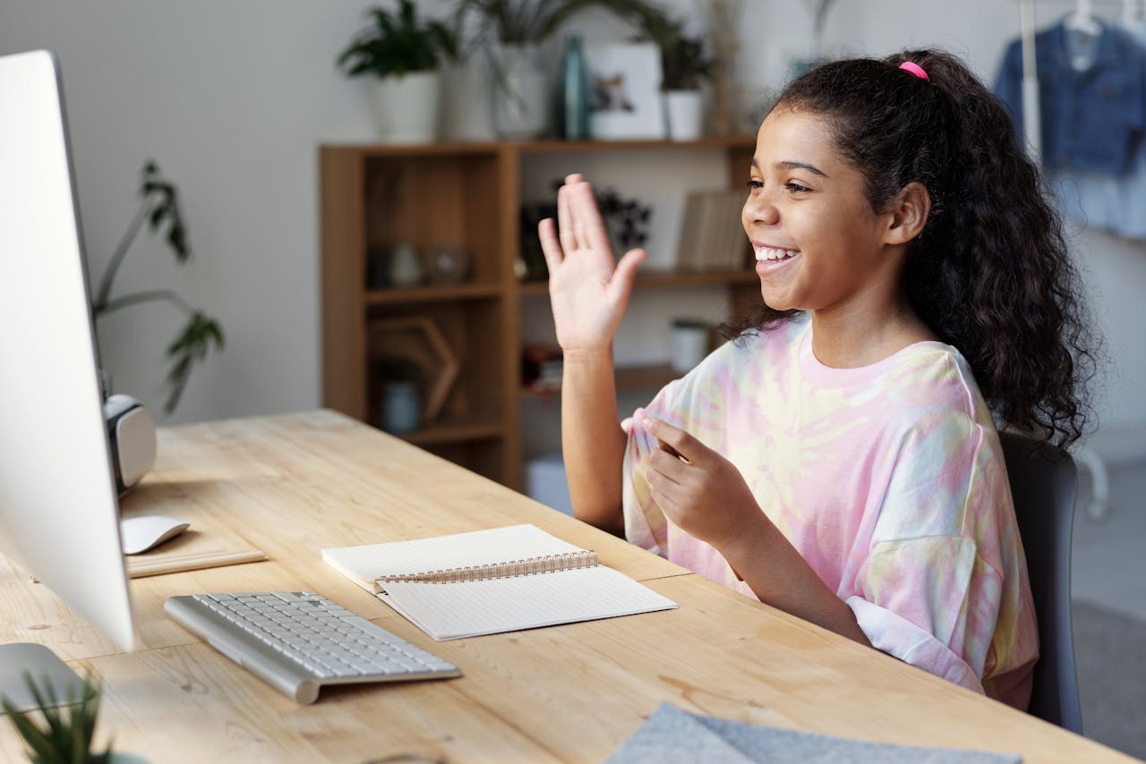 our-story A happy girl waves during an online class at home, sitting at a desk with a computer.